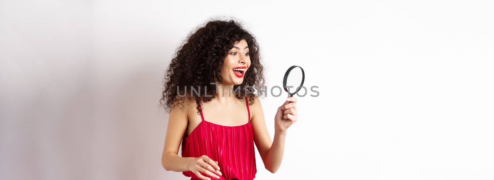 Excited woman in red dress look through magnifying glass and smiling, found interesting promo, standing on white background.