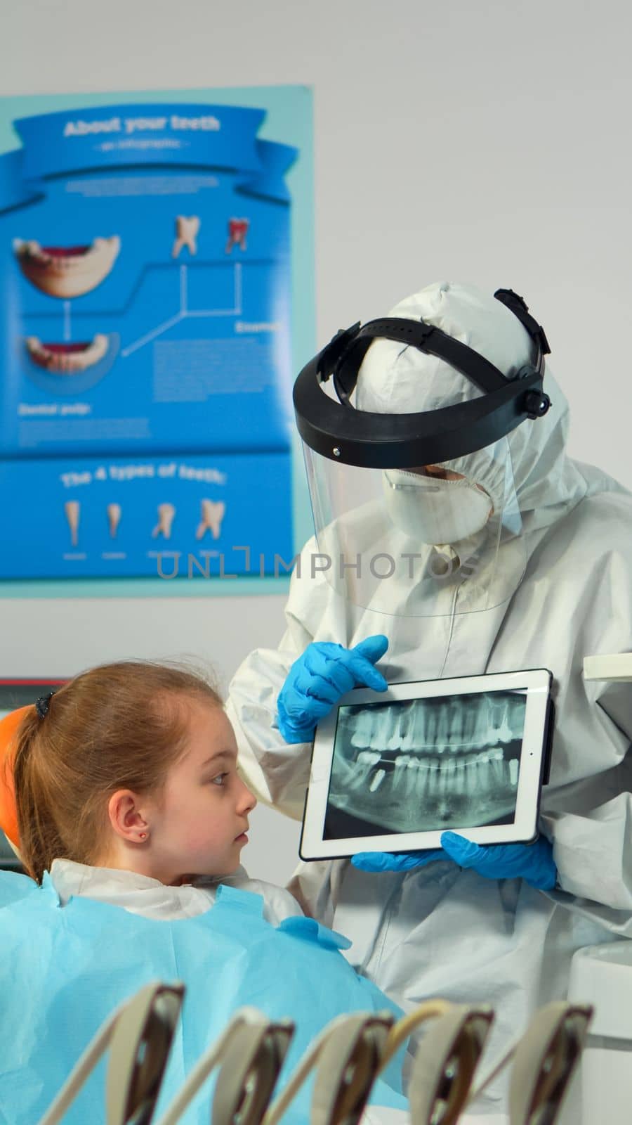 Dentist in protective equipment showing on tablet dental x-ray reviewing it with mother of kid patient. Medical team wearing face shield coverall, mask, gloves, explaining radiography using notebook