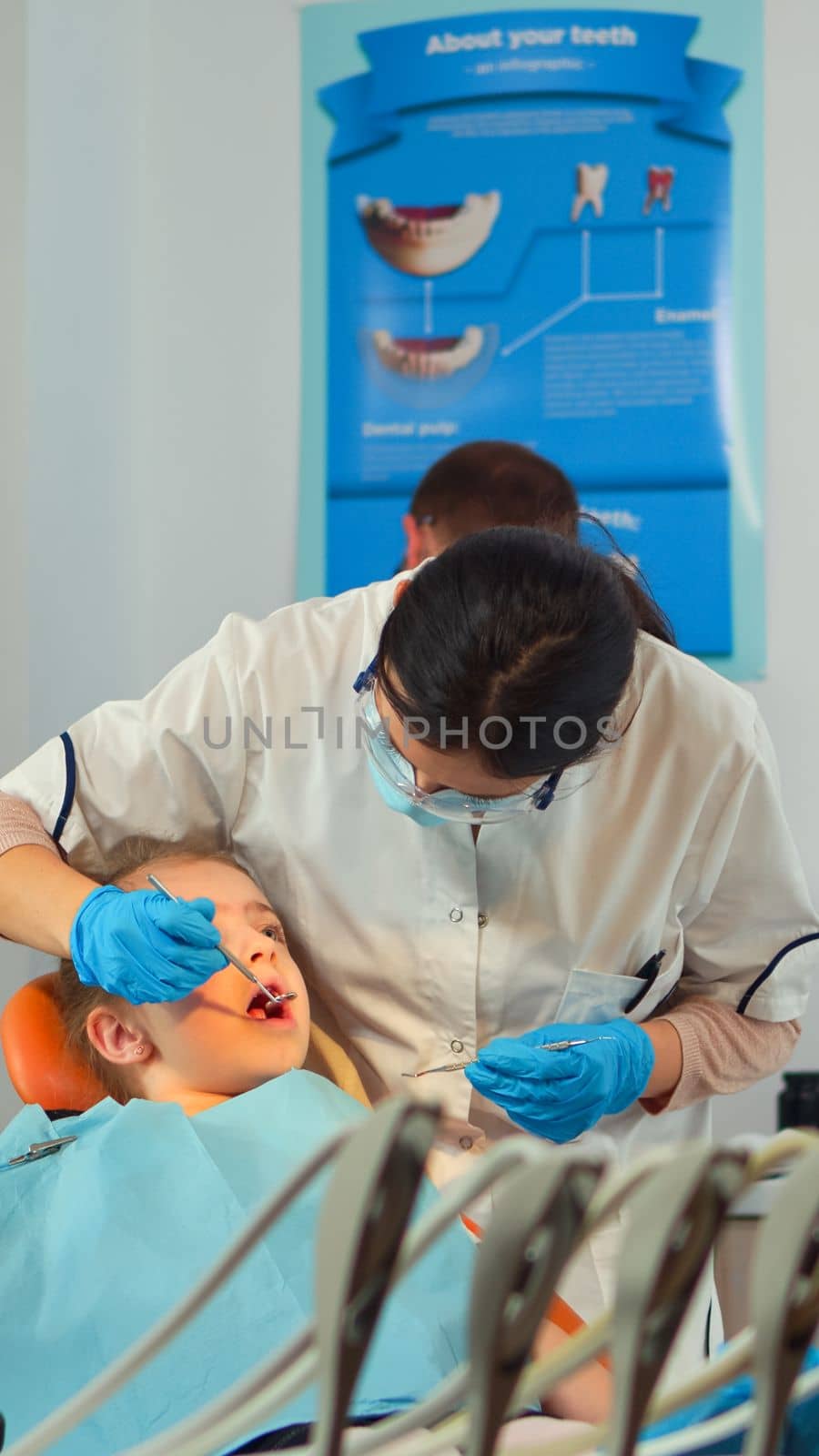 Stomatologist talking with mother of kids, lighting the lamp and examining little girl standing near stomatological chair. Pediatric dentist speaking to woman while nurse preparing sterilized tools.