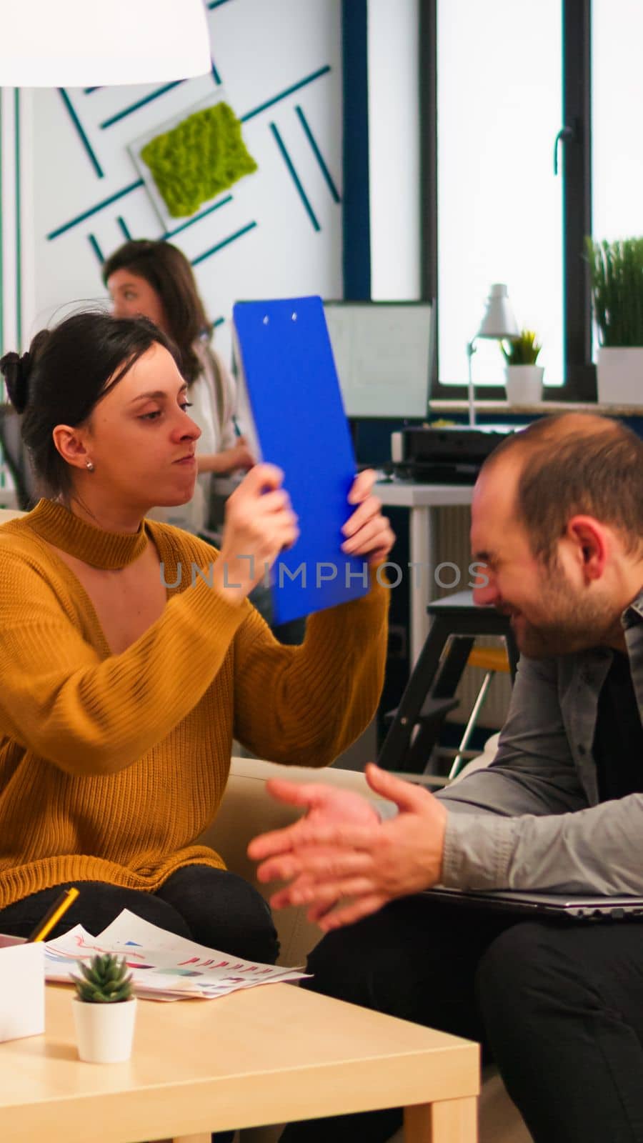 Angry manager woman screaming at diverse employees disagreeing about bad business contract, hitting man with clipboard in head sitting on couch at office desk, conflict dispute about document