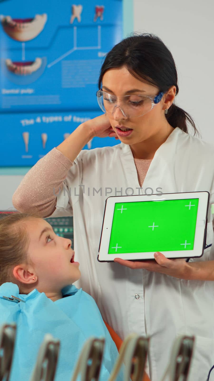 Dentist showing green screen display to mother of little patient while girl lying on dental chair. Woman explaining using monitor with green chroma key isolated chroma pc key mockup greenscreen