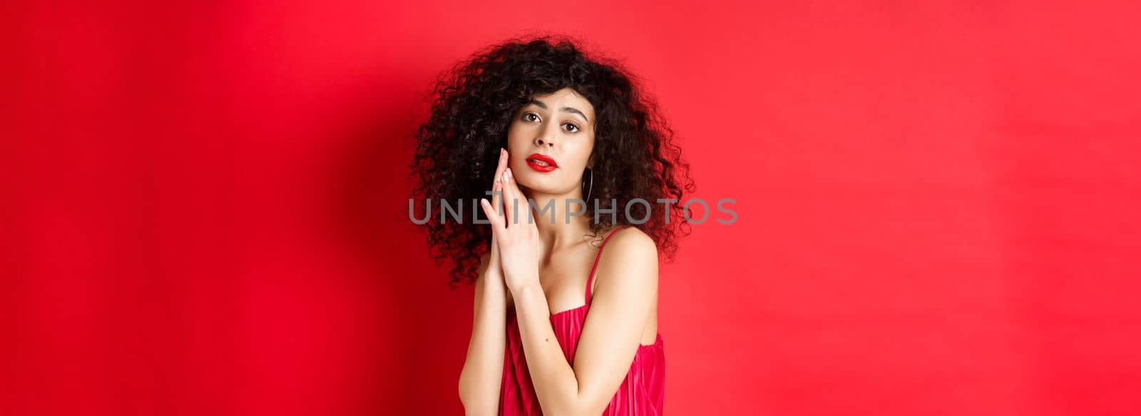 Romantic sensual woman with curly hair, wearing evening dress, posing seductive on red background by Benzoix