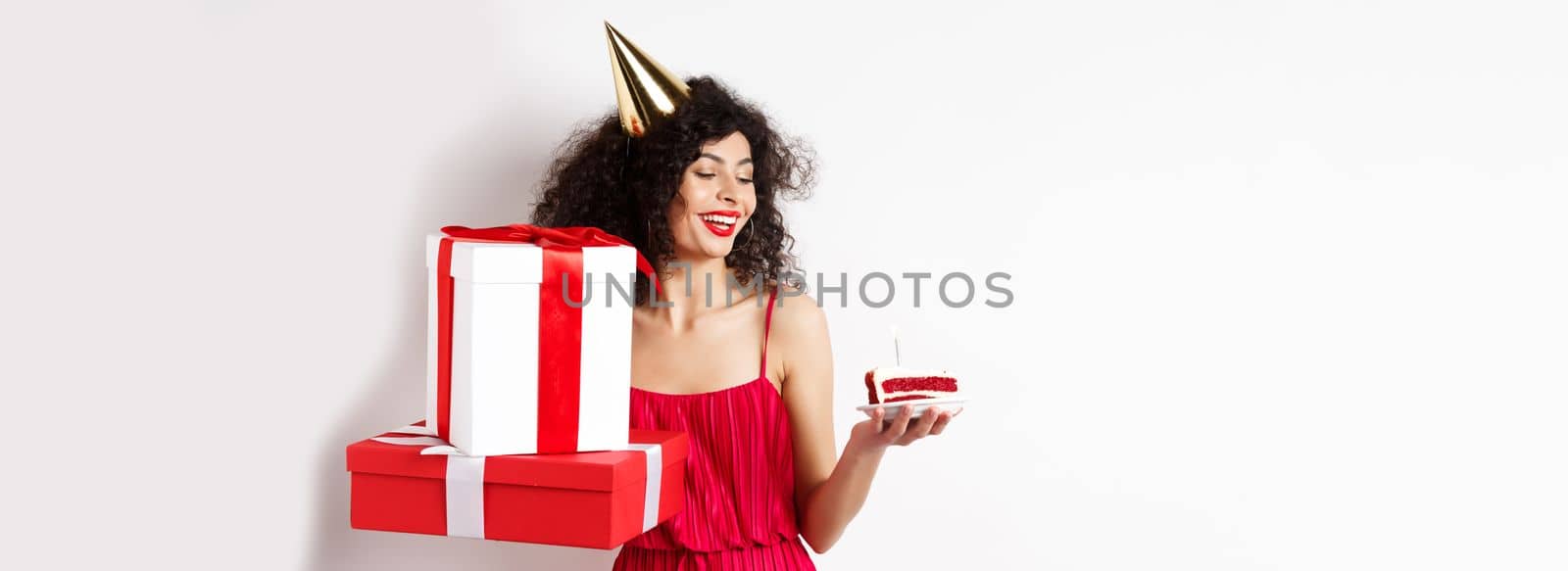 Cheerful smiling woman in red dress and party cone, looking happy at birthday cake, holding gifts, standing on white background. Copy space