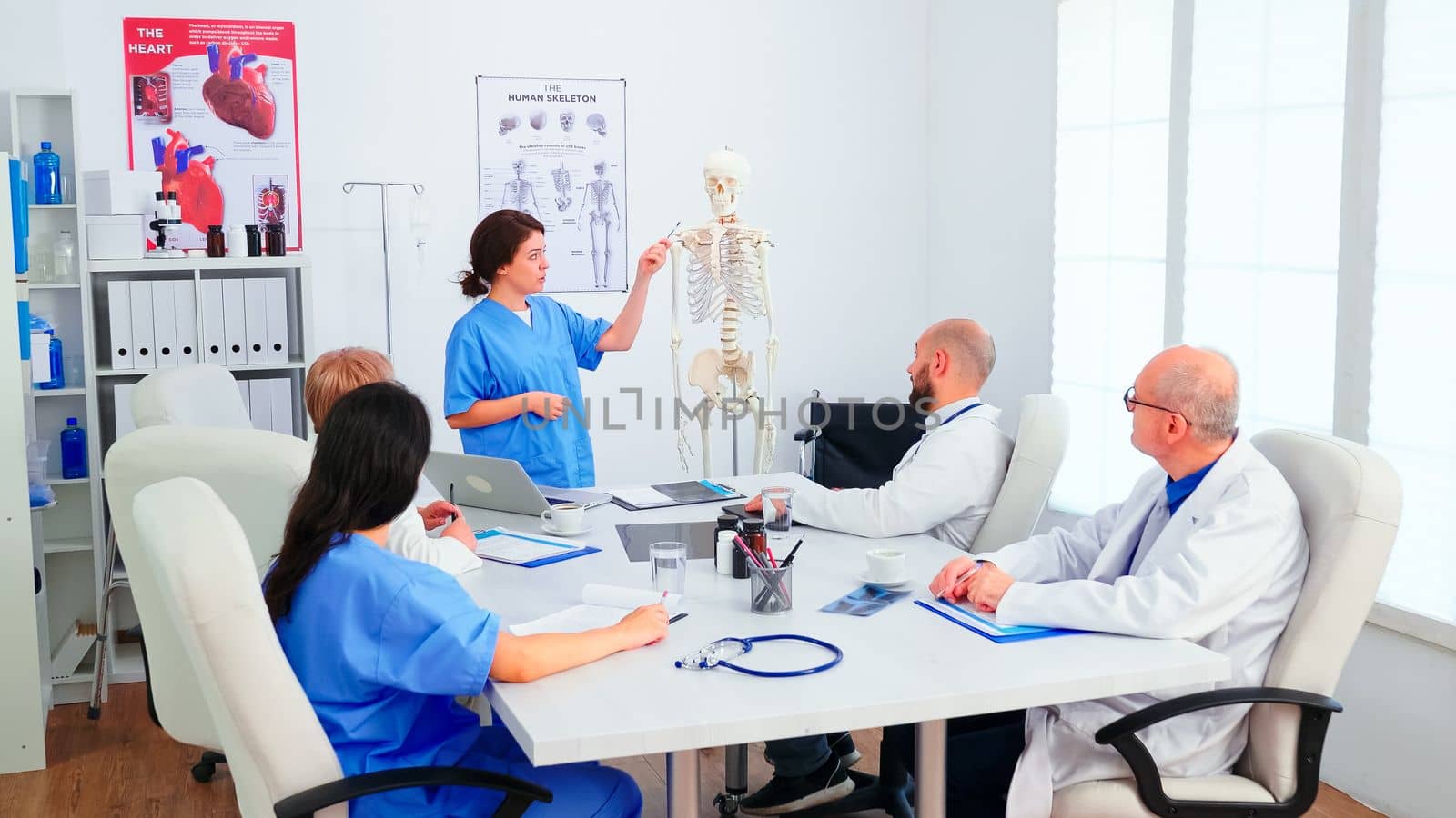 Young female assistent pointing on skeleton in front of medical surgeons in conference room. Clinic expert therapist talking with colleagues about disease, medicine professional