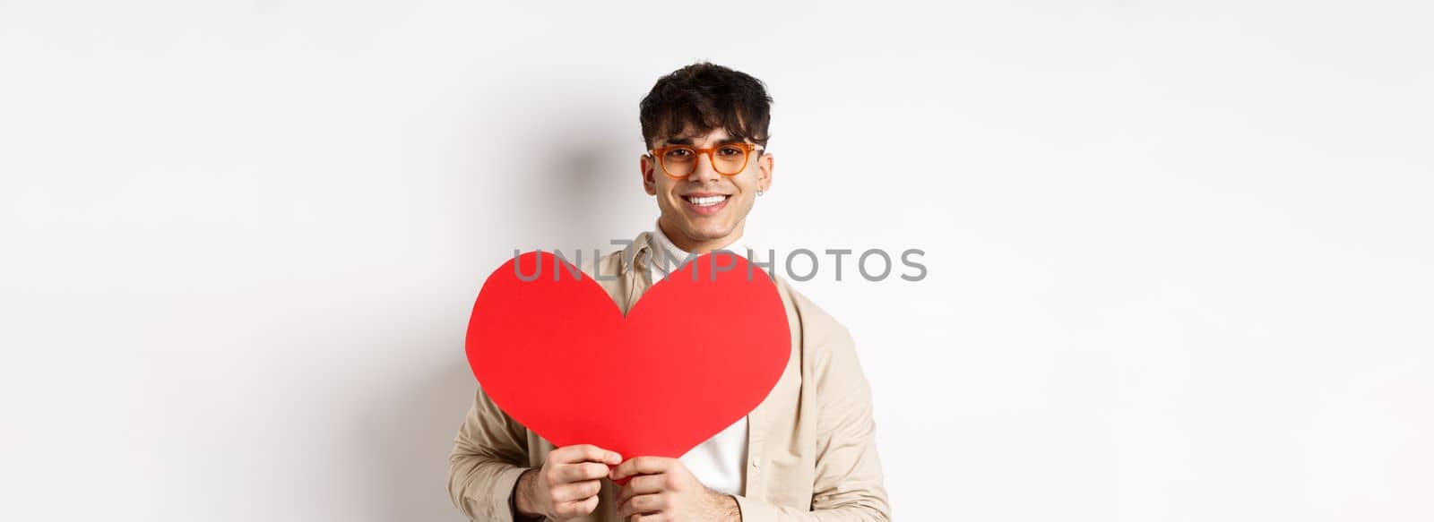 Handsome hipster guy with sunglasses and earring, waiting for true love on Valentines day, holding big red heart and smiling, standing over white background.