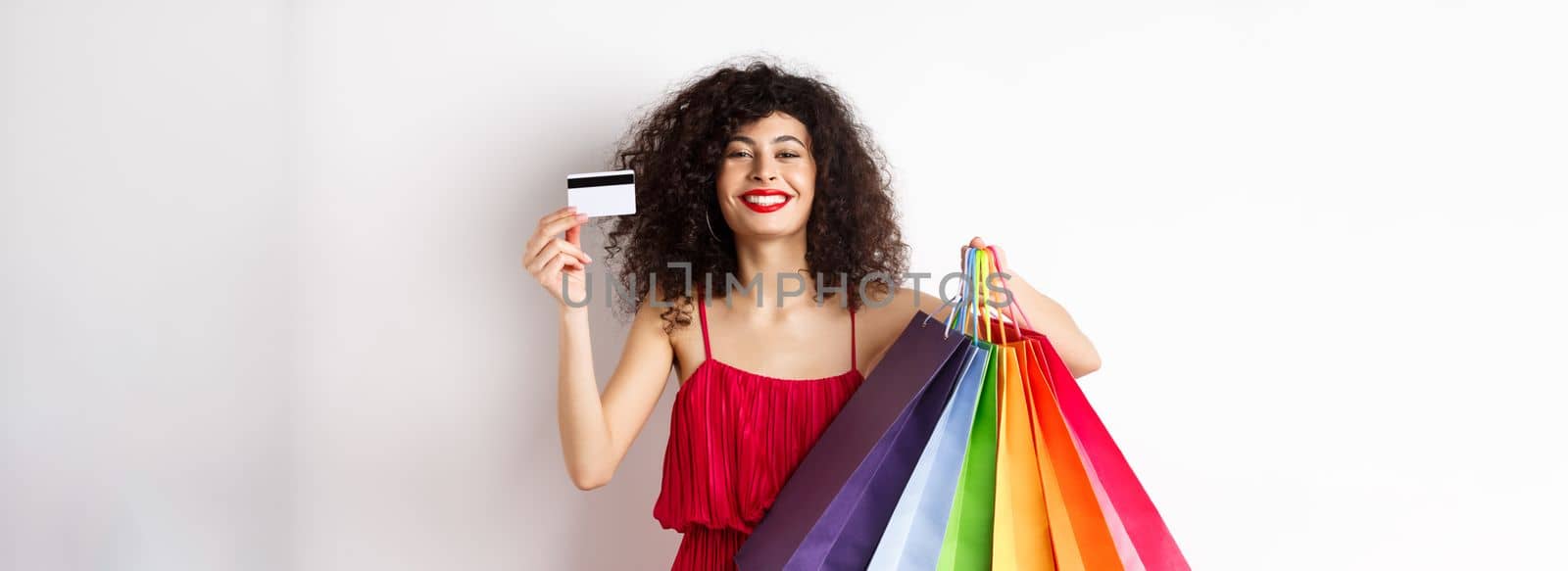 Happy elegant woman in red dress, showing shopping bags and plastic credit card, smiling pleased, standing on white background.