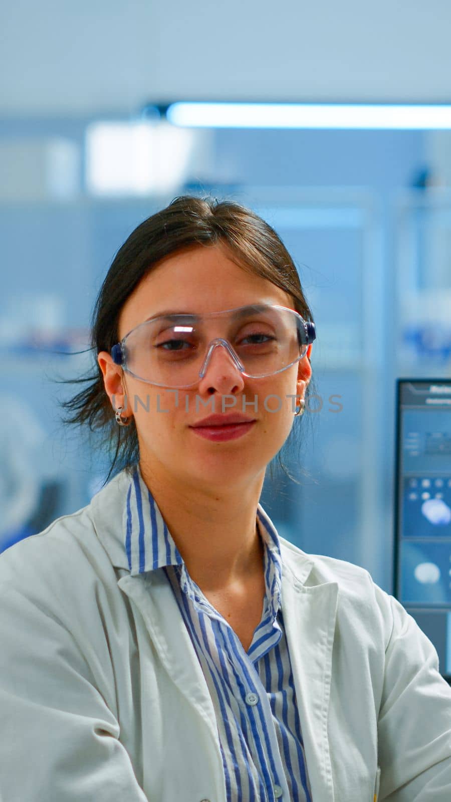 Chemist with lab coat sitting in laboratory looking at camera smiling, while colleague working on computer in background. Scientist examining virus evolution using high tech for scientific research
