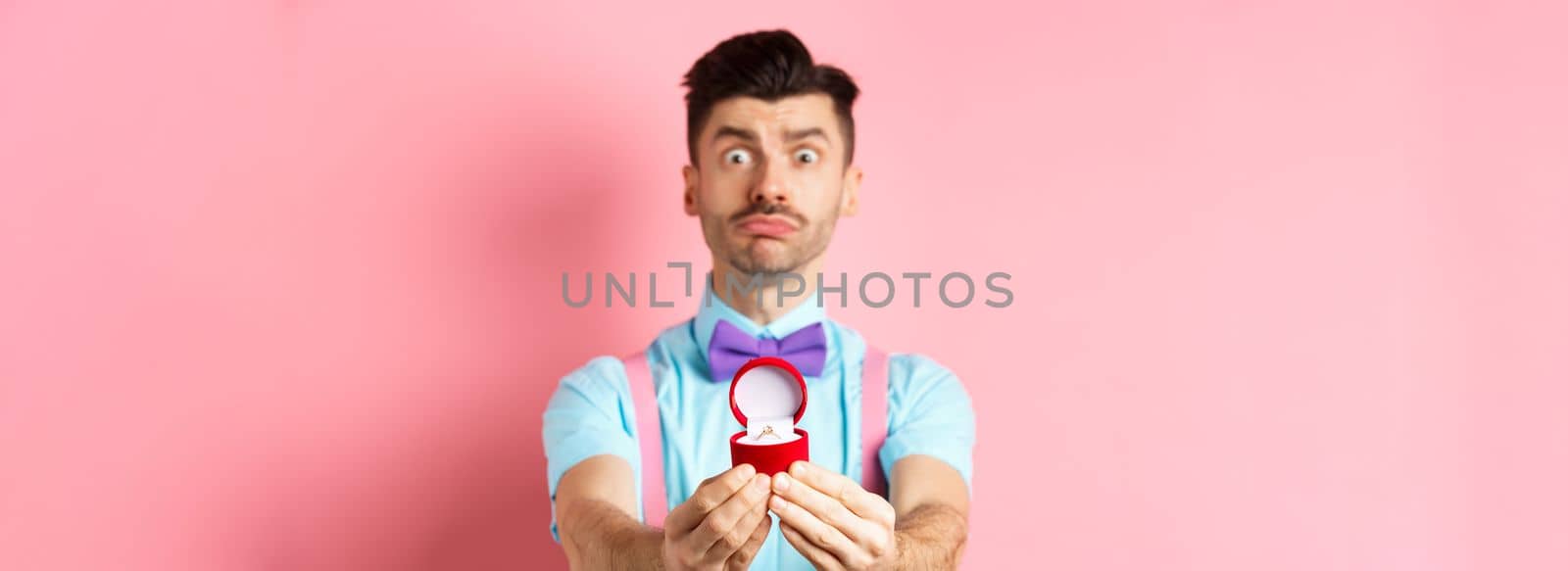 Valentines day. Nervous boyfriend waiting for girlfriend reply on marriage proposal, showing engagement ring and looking worried, standing over pink background.