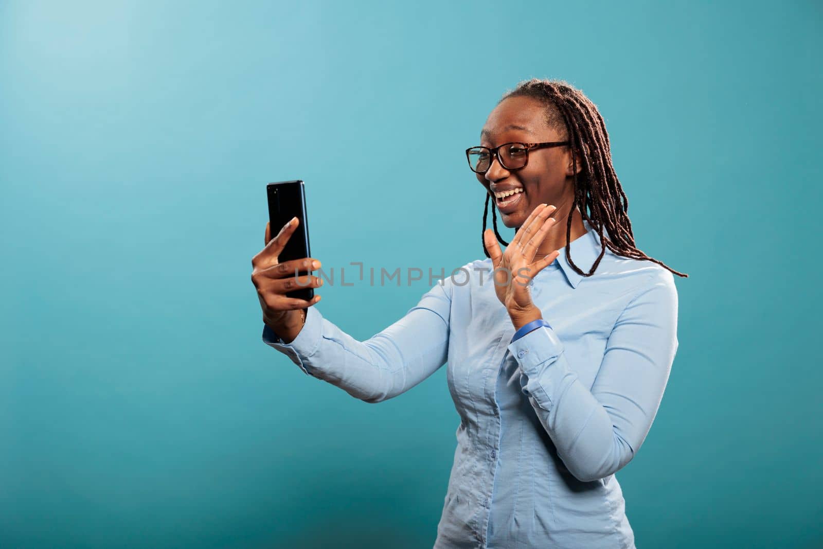 Happy smiling african american woman with smartphone waving at video conference call on blue background. Beautiful confident young lady greeting friend over online phone video call.
