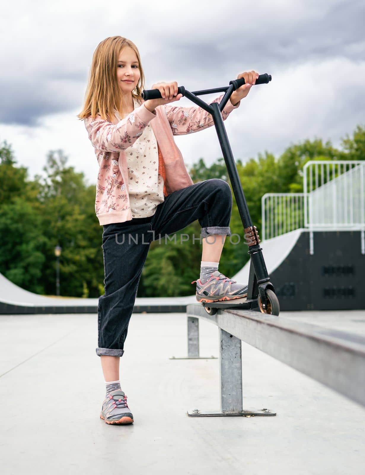 Preteen girl standing with scooter at street in the park and looking at camera. Cute child posing with eco vehicle outdoors