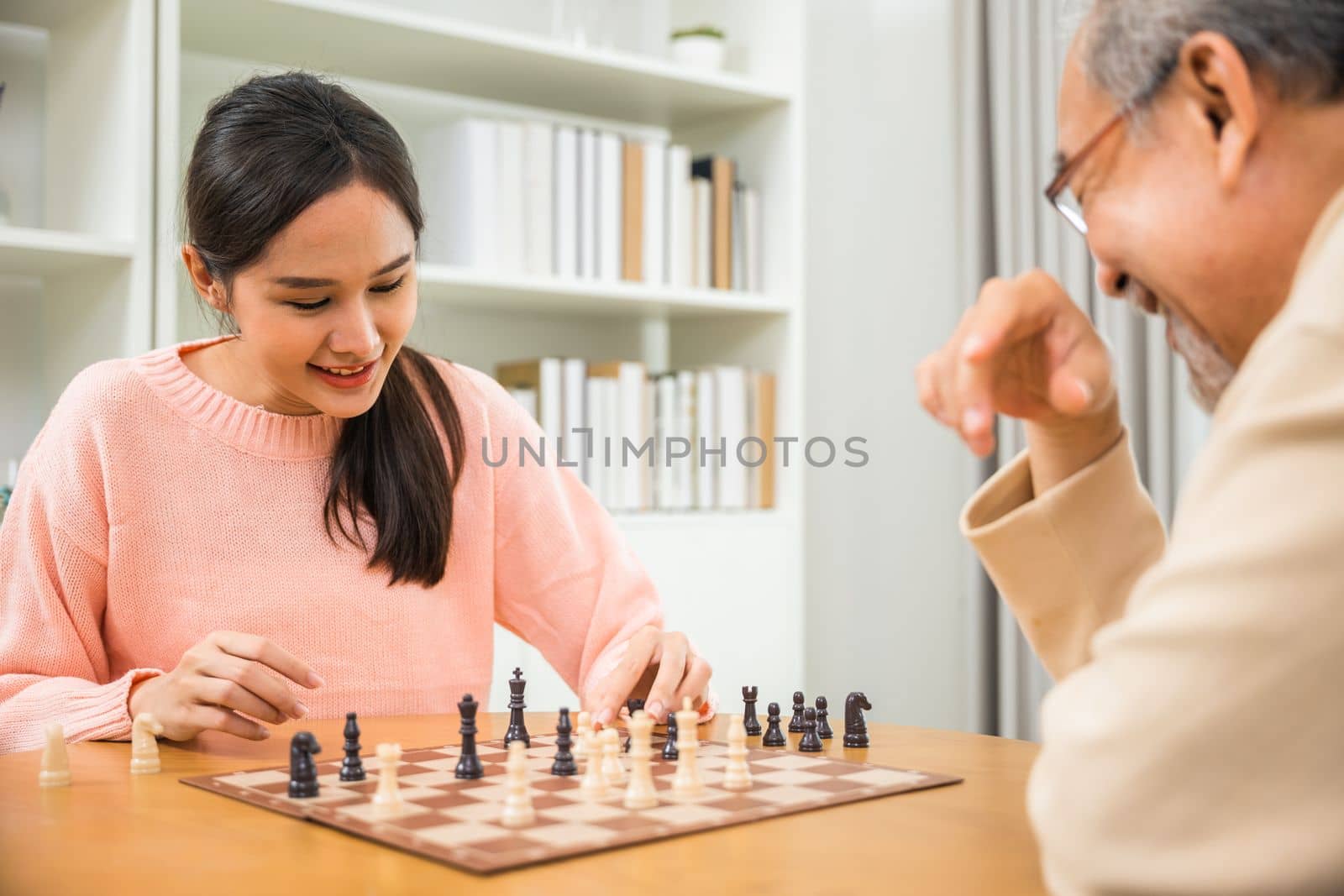 Beautiful young smile woman having fun sitting playing chess game with senior elderly at home by Sorapop