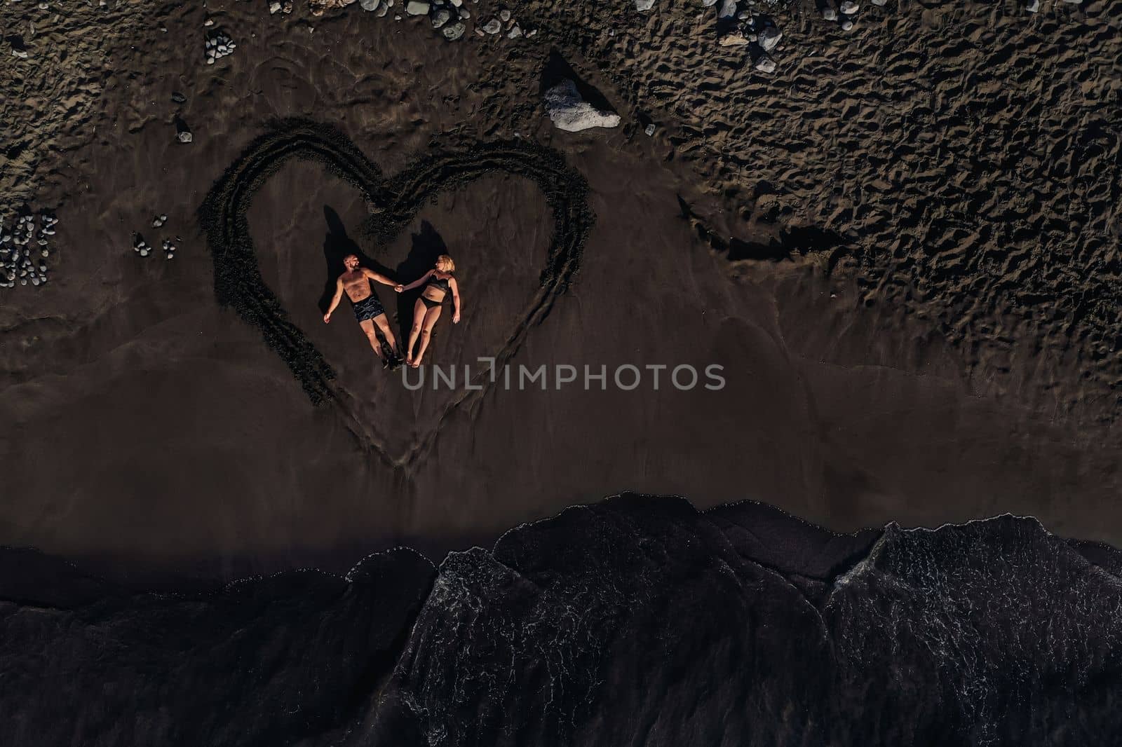 A couple in love lies on a beach of black volcanic sand in a painted heart, Tenerife, Canary Islands, Spain.
