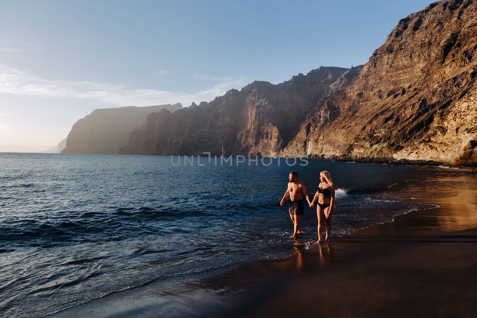 A couple in love walks on the beach against the backdrop of the Acantilados de Los Gigantes mountains at sunset, Tenerife, Canary Islands, Spain.