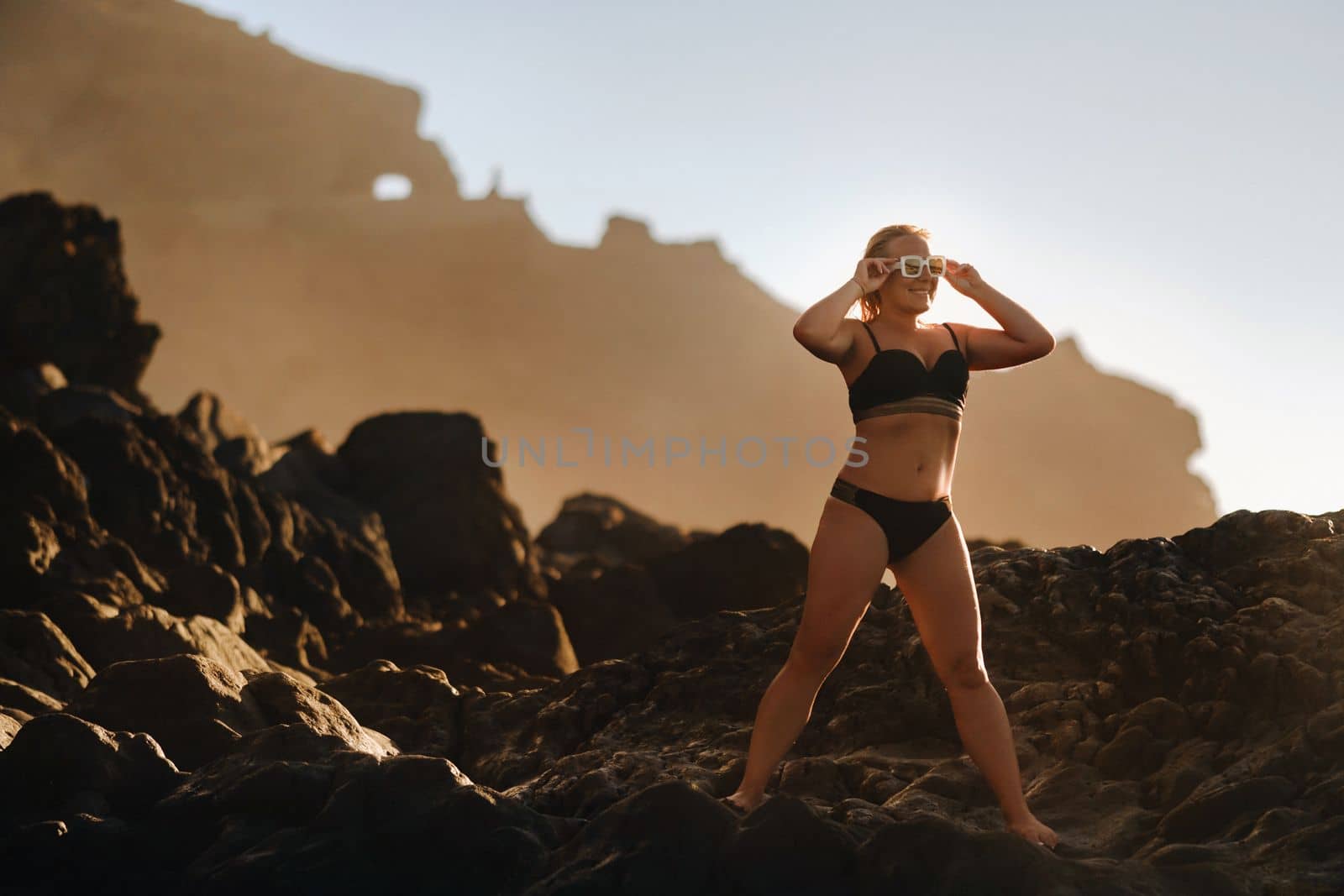 a girl in a black swimsuit and glasses stands on the rocks near the beach by the sea in summer at sunset.
