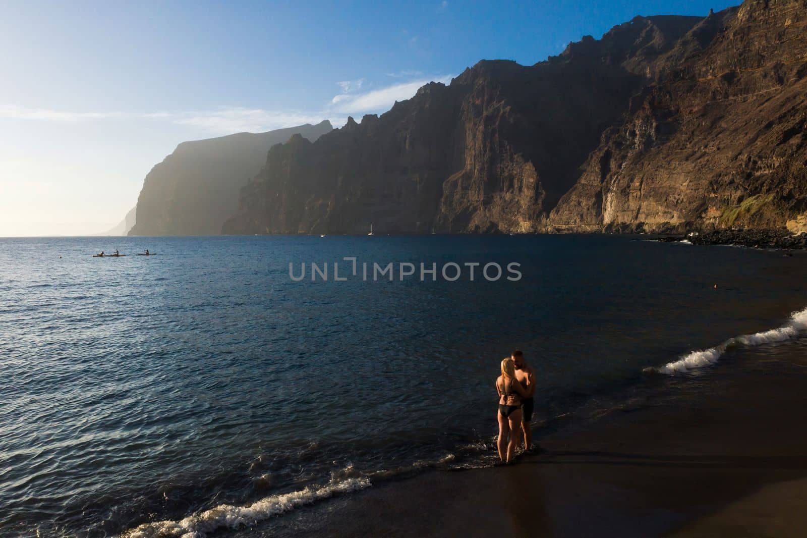 A couple in love stands on a black beach near the rocks of Acantilados de Los Gigantes at sunset, Tenerife, Canary Islands, Spain by Lobachad