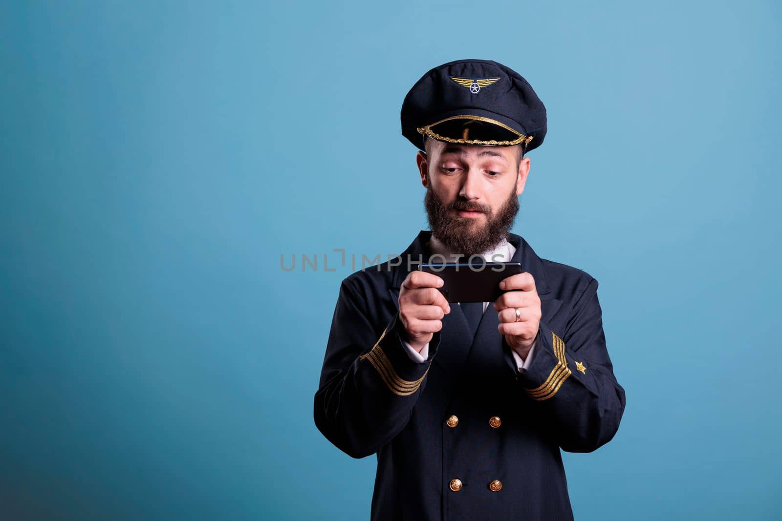 Plane pilot in professional uniform playing mobile games on smartphone, holding phone, watching online video. Airlane captain using aviation simulator on telephone, leisure entertainment