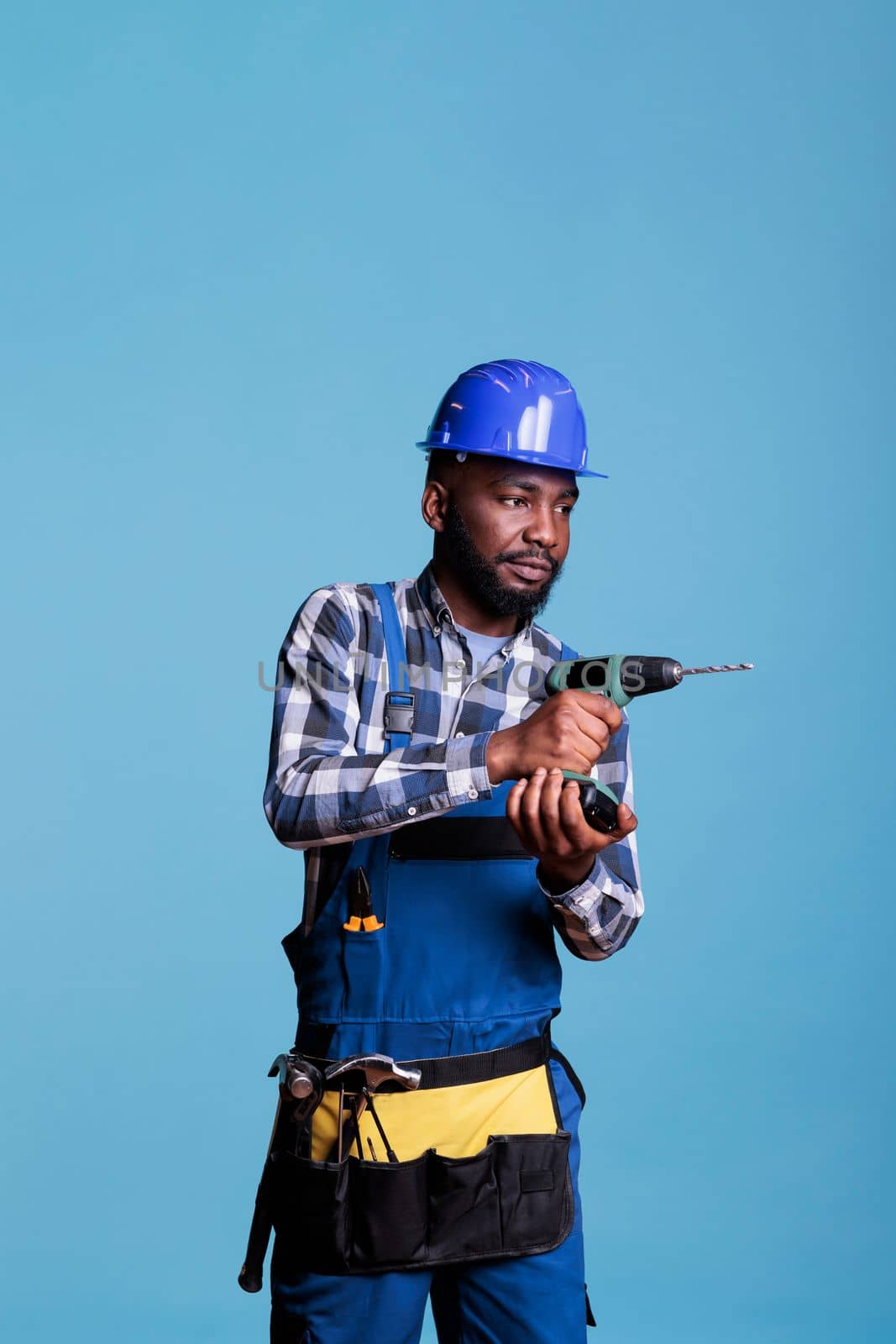 African american builder with coverall drilling a wall for a building renovation inside. Builder wearing hard hat insulated on blue background in studio, construction industry concept.