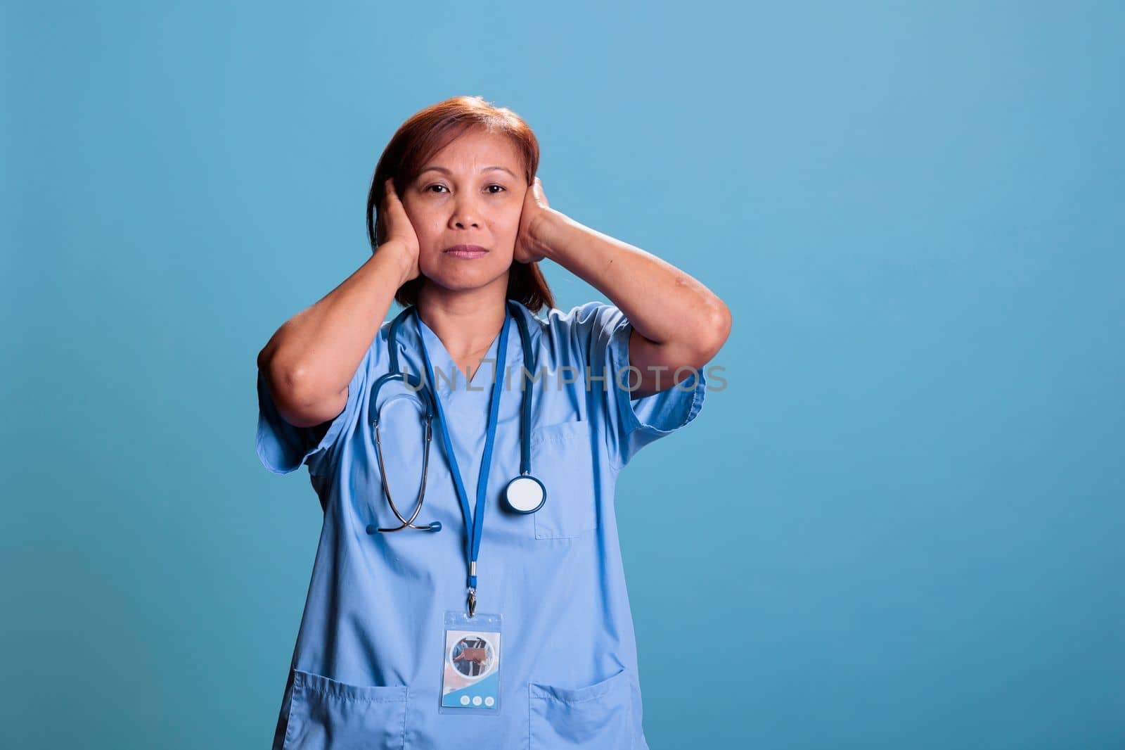 Asian quiet nurse with stethoscope covering ears having serious expression by DCStudio