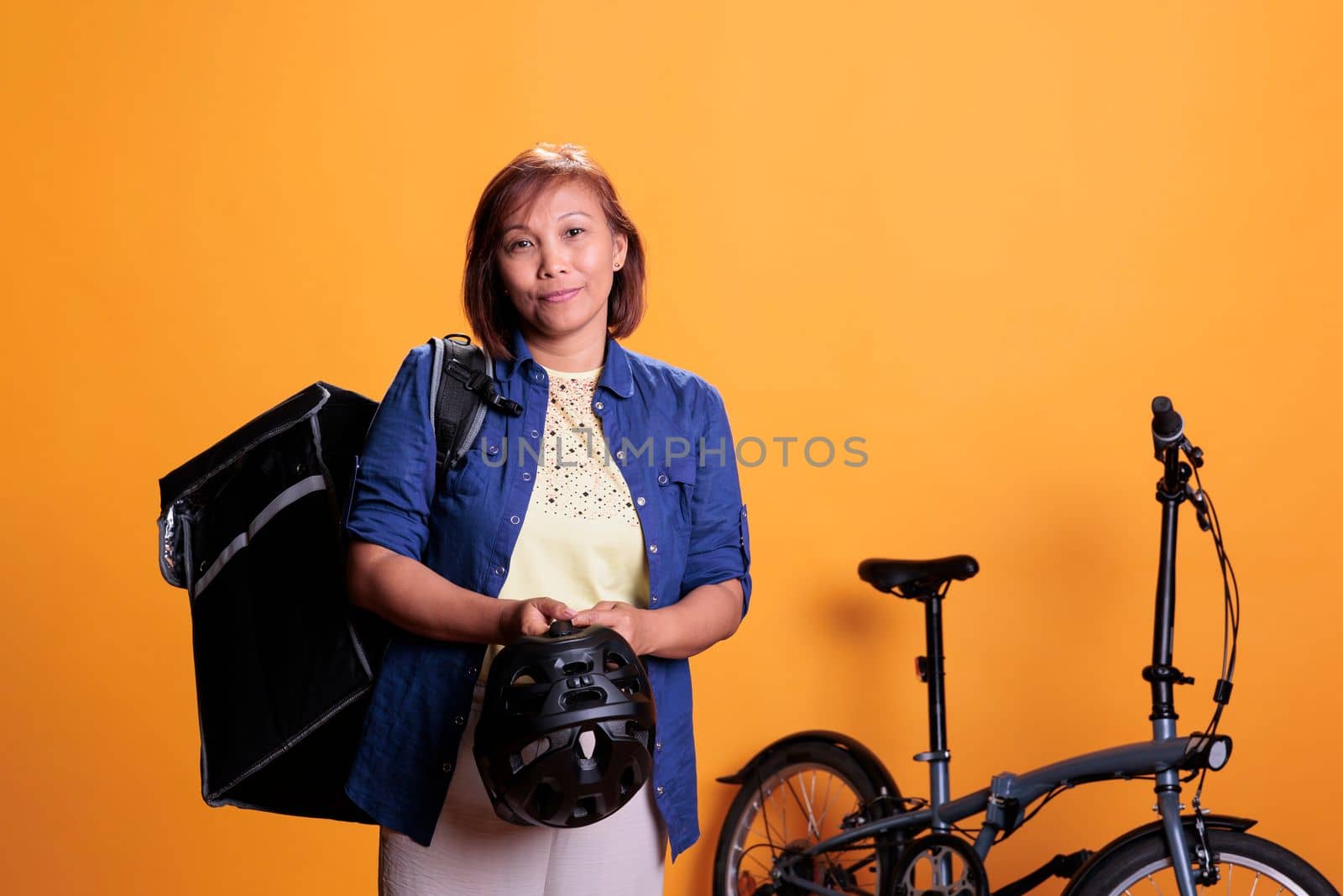 Elderly deliverywoman carrying thermal delivery backpack standing beside bike in studio with yellow background. Restaurant worker delivering pizza for lunch. Food transportation service and concept