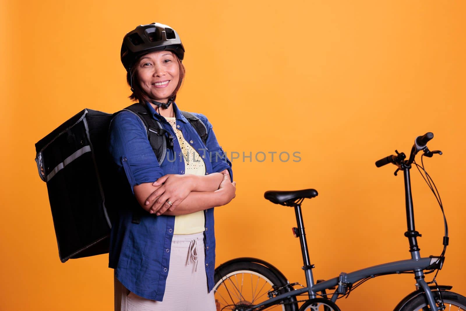 Asian deliver carrying food takeaway backpack standing beside bike in studio with yellow background. Restaurant courier delivering pizza for dinner. Food transportation service and takeaway concept