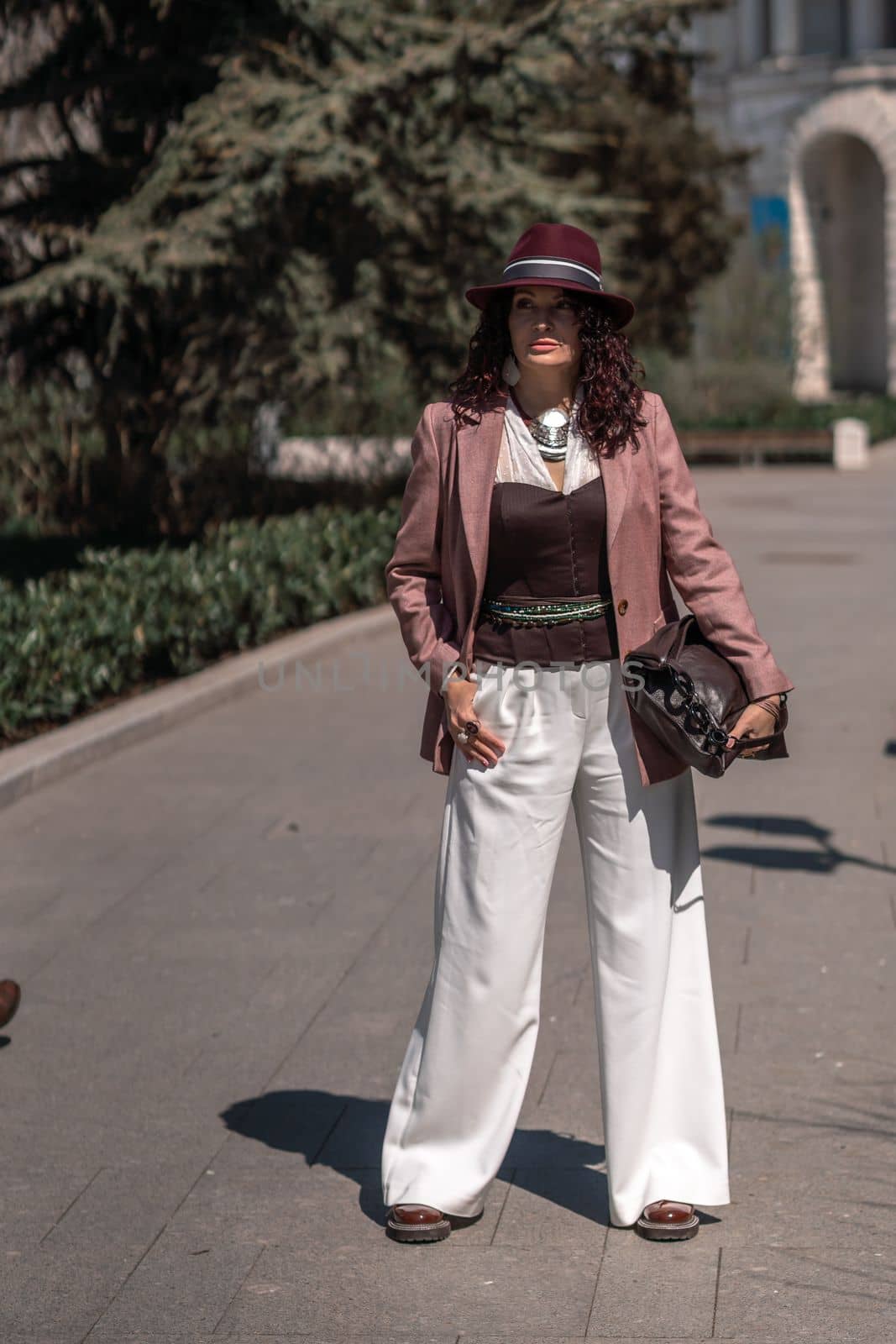 Woman park city. Stylish woman in a hat walks in a park in the city. Dressed in white corset trousers and a pink jacket with a bag in her hands