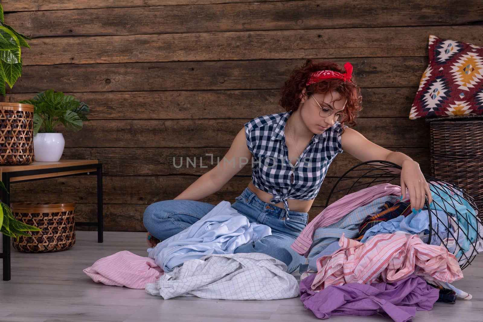 Attractive girl sorts clothes for washing. Short curly hair. Metal basket full of clothes prepared for washing. A wall of brown boards.