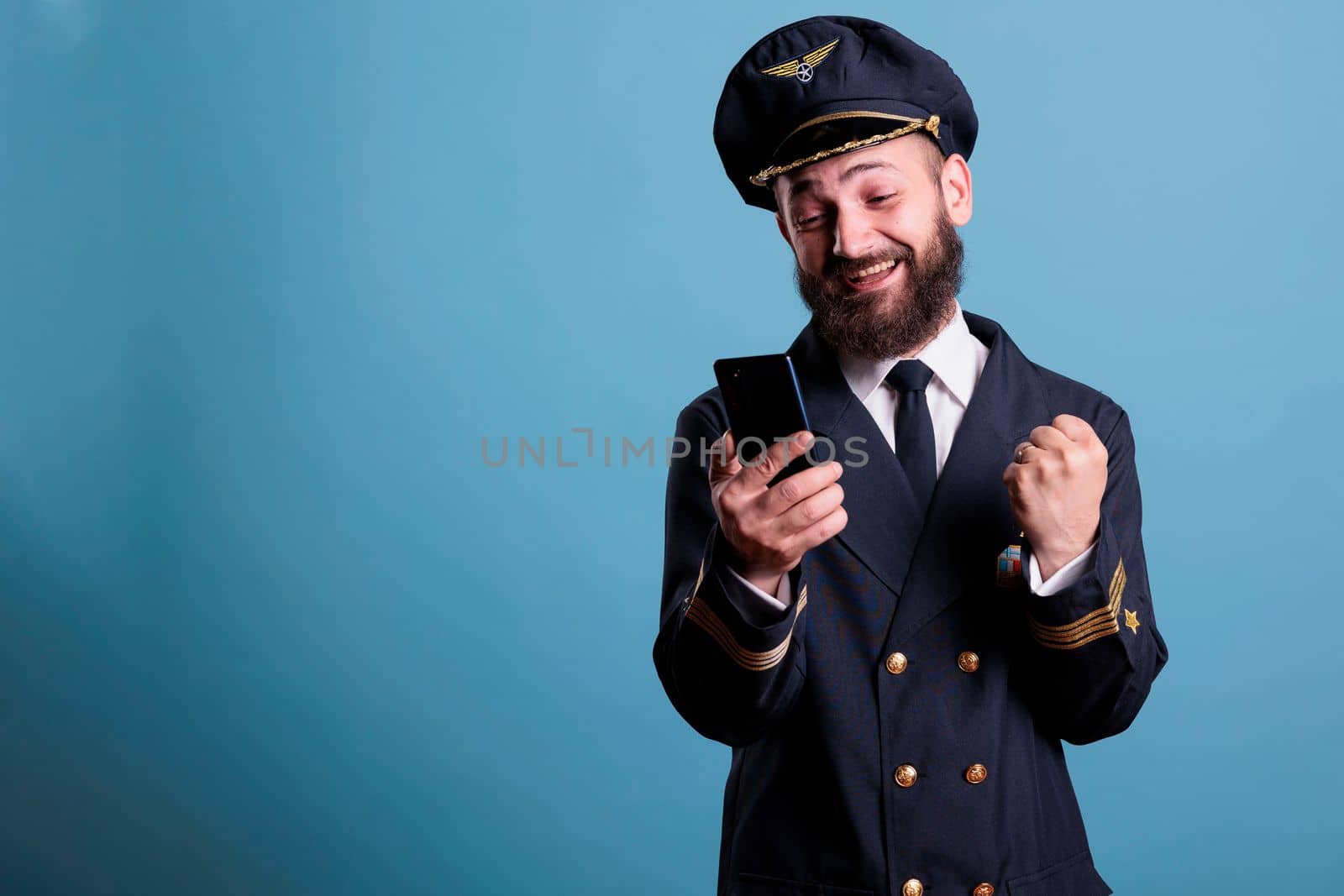 Happy excited airplane aviator in uniform holding smartphone, making winner gesture, clenched fist. Pilot with cheerful facial expression looking at mobile phone screen, reading good news