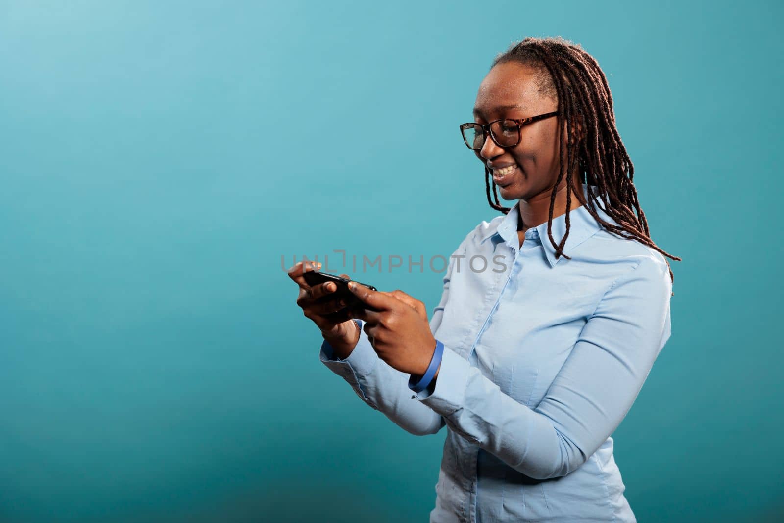 Confident joyful young adult person with modern cellphone watching video content. Beautiful happy woman with touchscreen smartphone browsing internet webpages while standing on blue background.