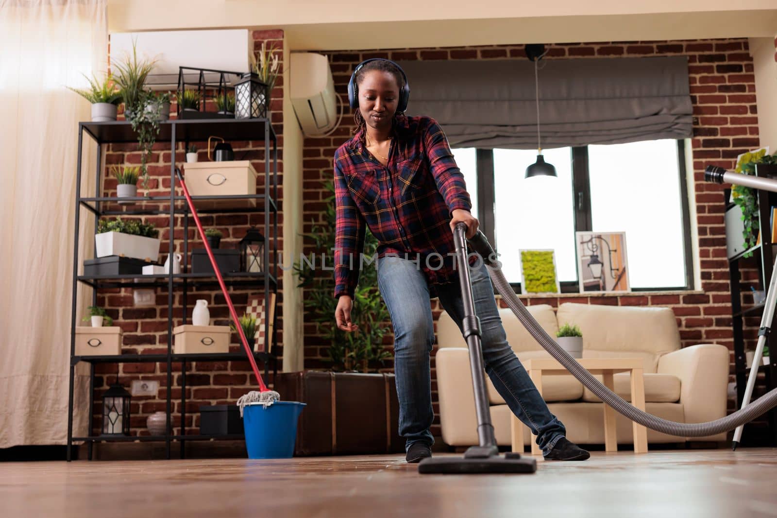 Positive housewife doing spring cleaning at home, singing while vacuuming in living room. Listening to music with wireless headset relaxed looking cheerful, taking care of finishing household chores.