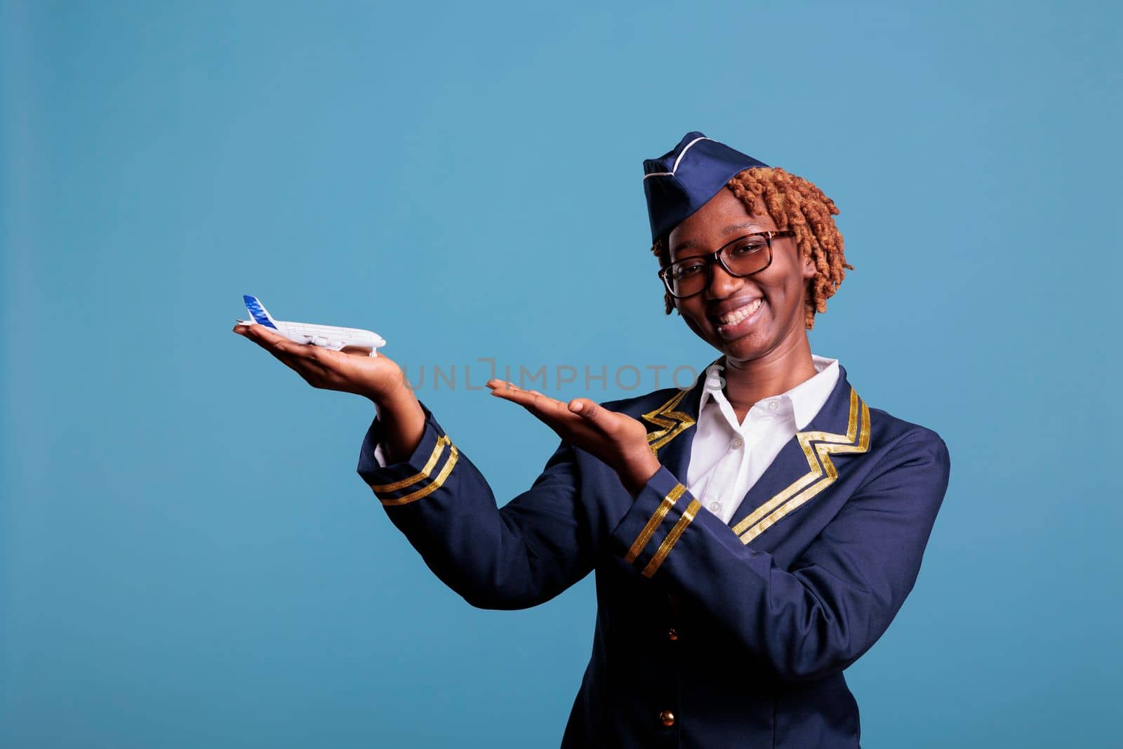 Smiling african american stewardess pointing at commercial airplane holding in hand for airline advertising. Flight attendant wearing crew members uniform in studio shot.