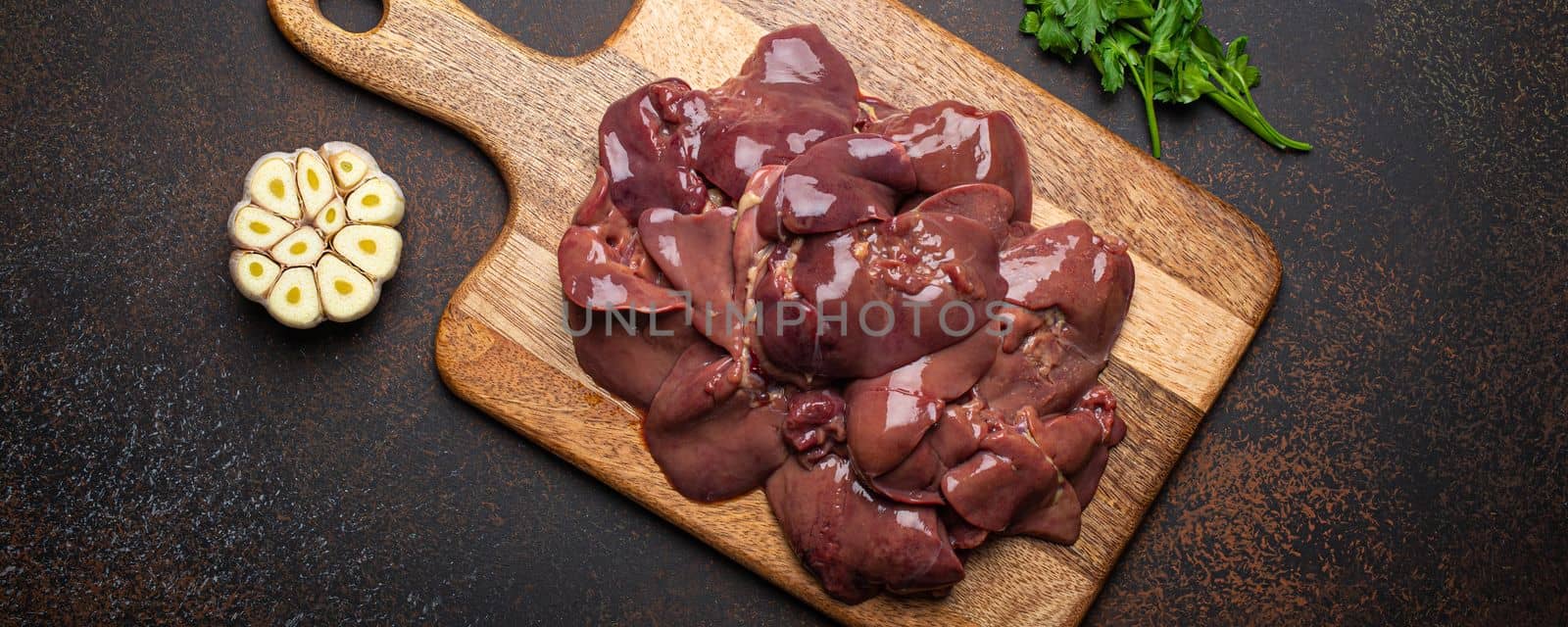 Raw chicken liver on wooden cutting board top view on dark rustic concrete background kitchen table with parsley and garlic. Healthy food ingredient, source of iron, folate, vitamins and minerals