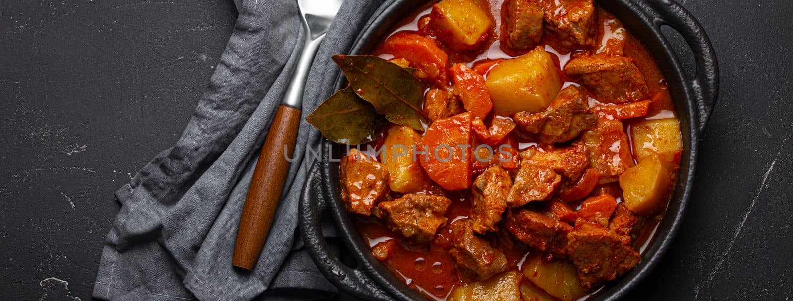 Beef meat stew with potatoes, carrot and delicious gravy in black casserole pot with bay leaves with spoon on black dark rustic concrete background from above .