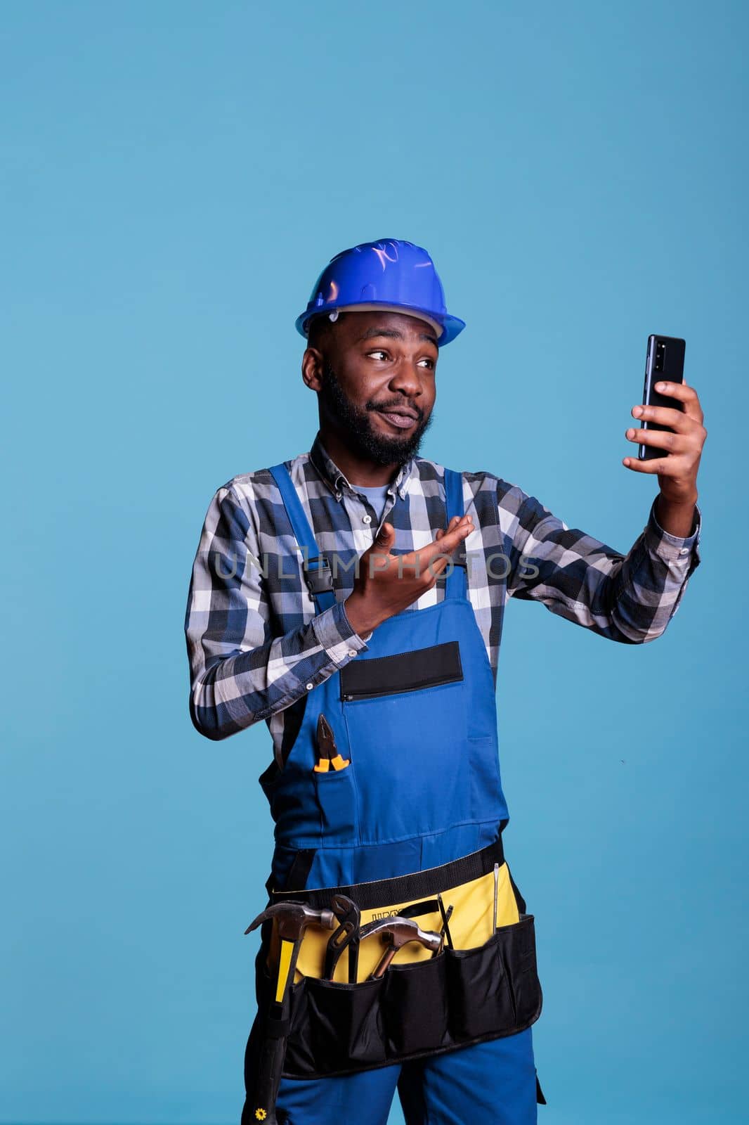 African american contractor talking on video call with business partner wearing coveralls against blue background in studio shot. Construction worker talking pleasantly on cell phone, wearing hard hat and tool belt.