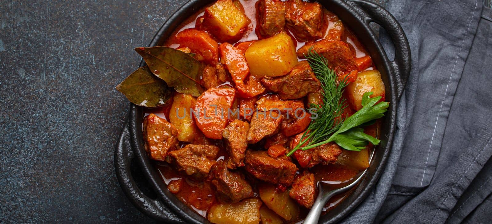 Beef meat stew with potatoes, carrot and delicious gravy in black casserole pot with bay leaves and fresh green herbs with spoon on black dark rustic concrete background from above . by its_al_dente