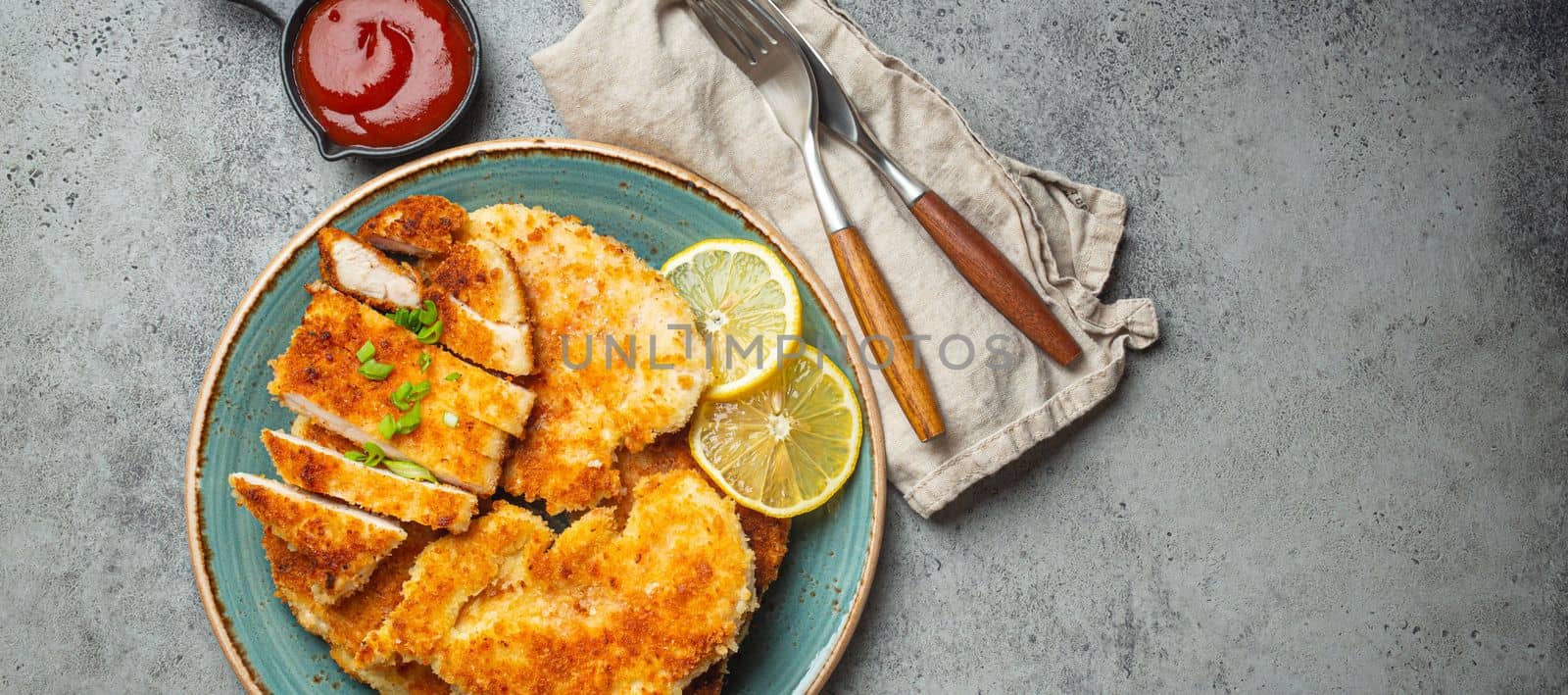 Crispy panko breaded fried chicken fillet with green salad and lemon cut on plate on gray background table with ketchup from above. Japanese style deep fried coated chicken breasts, space for text.
