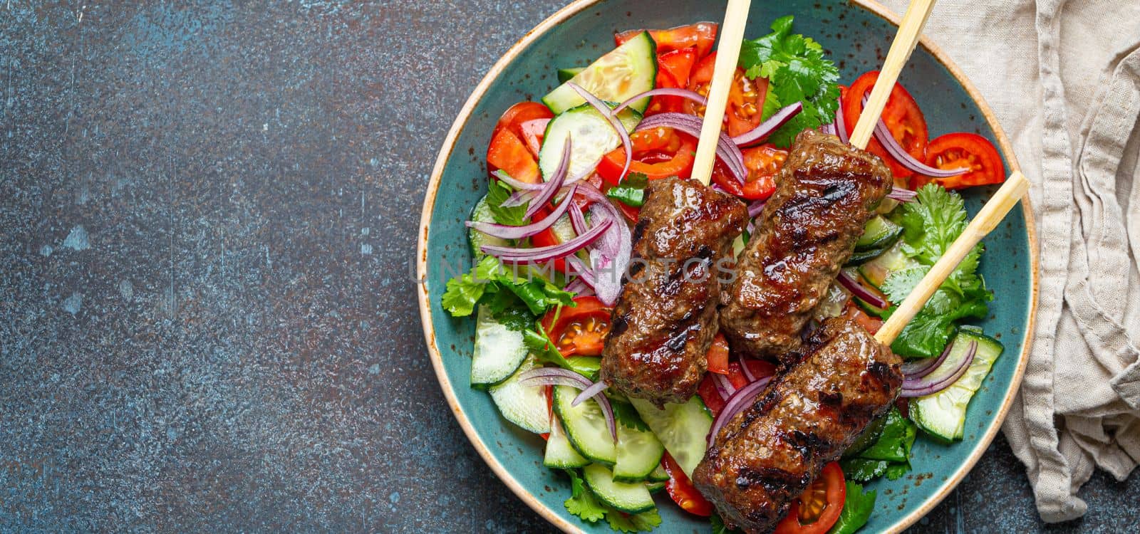 Grilled skewer meat beef kebabs on sticks served with fresh vegetables salad on plate on rustic concrete background from above. Traditional Middle Eastern Turkish dish Kebab, space for text by its_al_dente