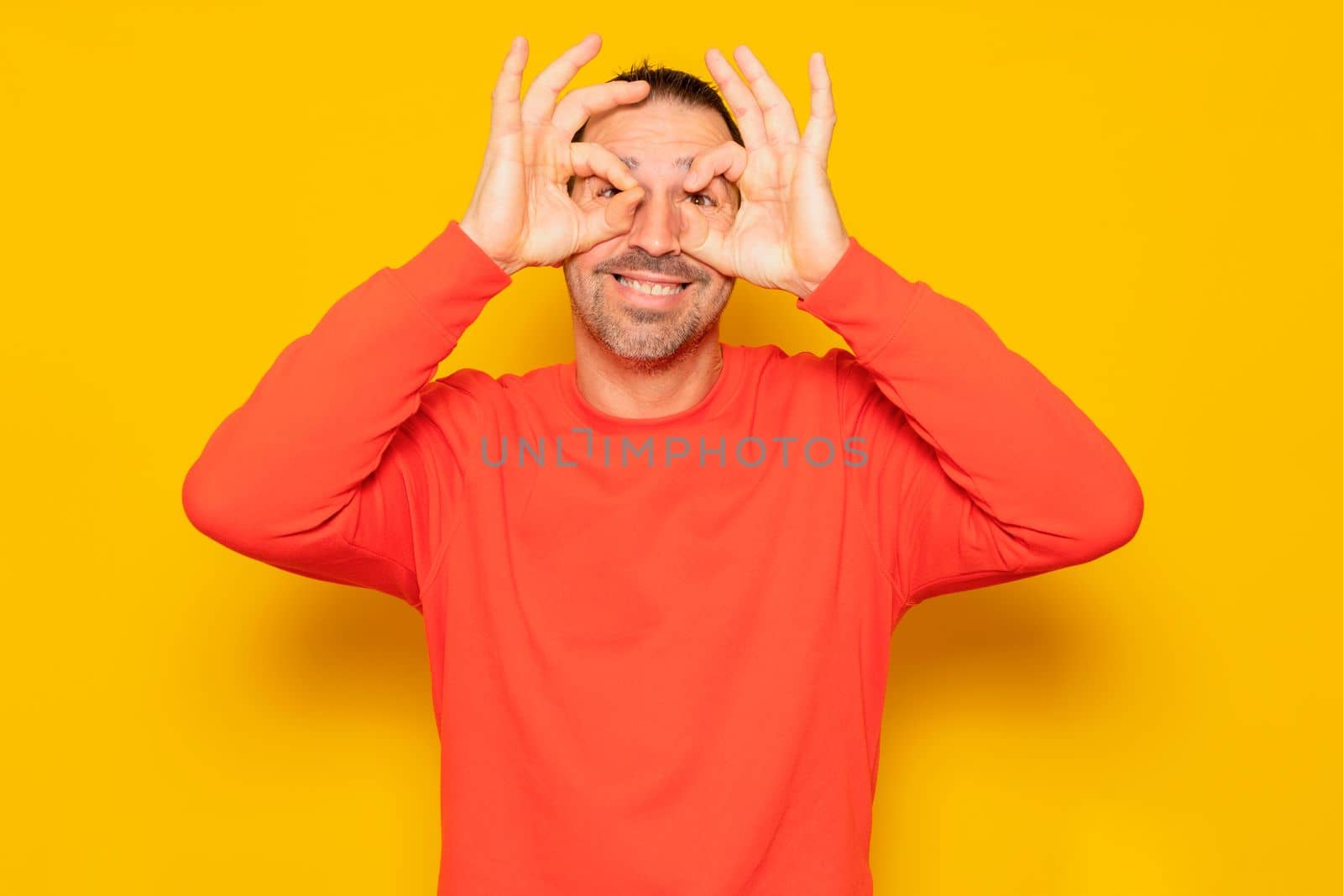 Handsome hispanic man with beard wearing red jumper over isolated orange background doing ok gesture like binoculars, eyes looking through ok shaped fingers. Crazy expression