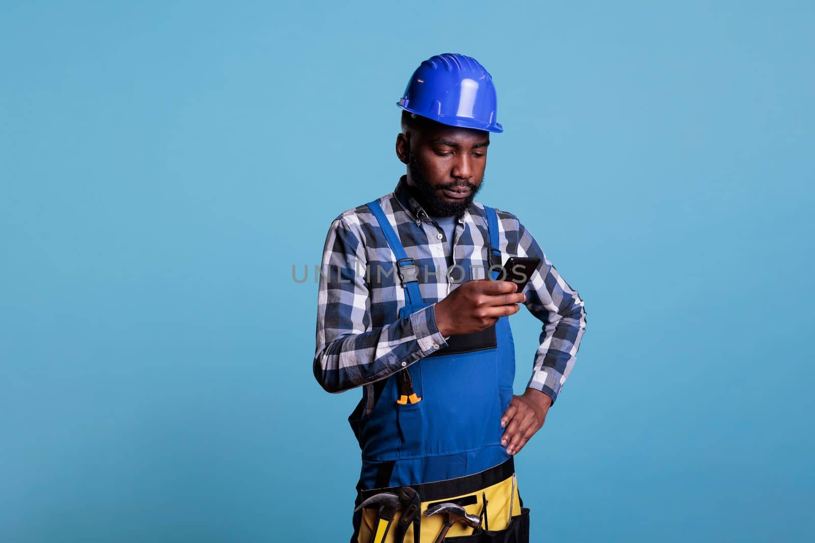 Distracted african american construction worker using cell phone in studio shot against blue background. Contractor wearing work uniform and hard hat while texting with mobile device.