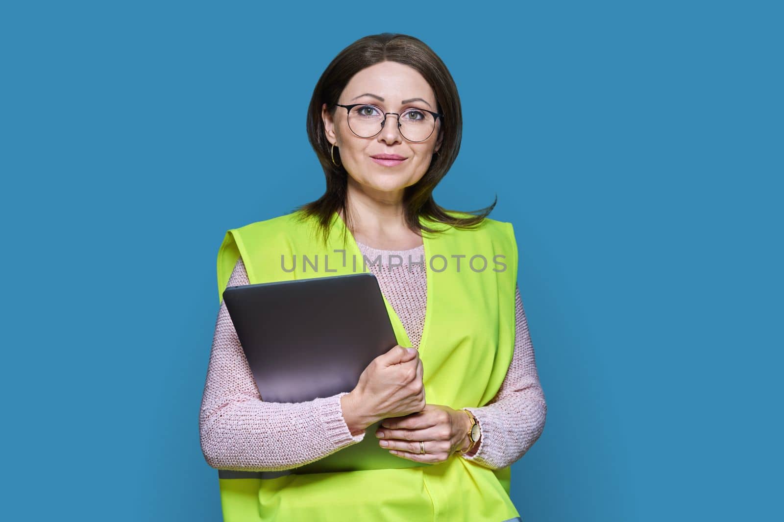 Portrait of engineer manager woman in helmet vest holding laptop, over blue studio background. Logistics, construction, industry, management, architecture, engineering, staff workers concept