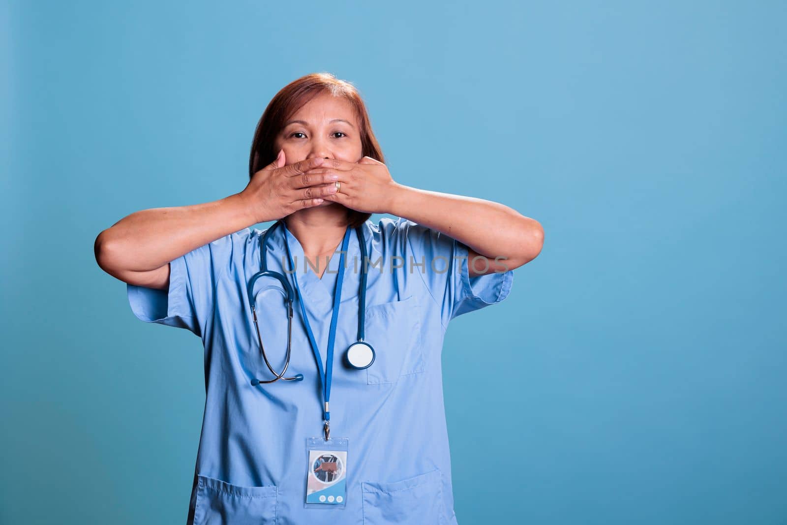 Elderly physician nurse in blue uniform covering mouth with hands by DCStudio