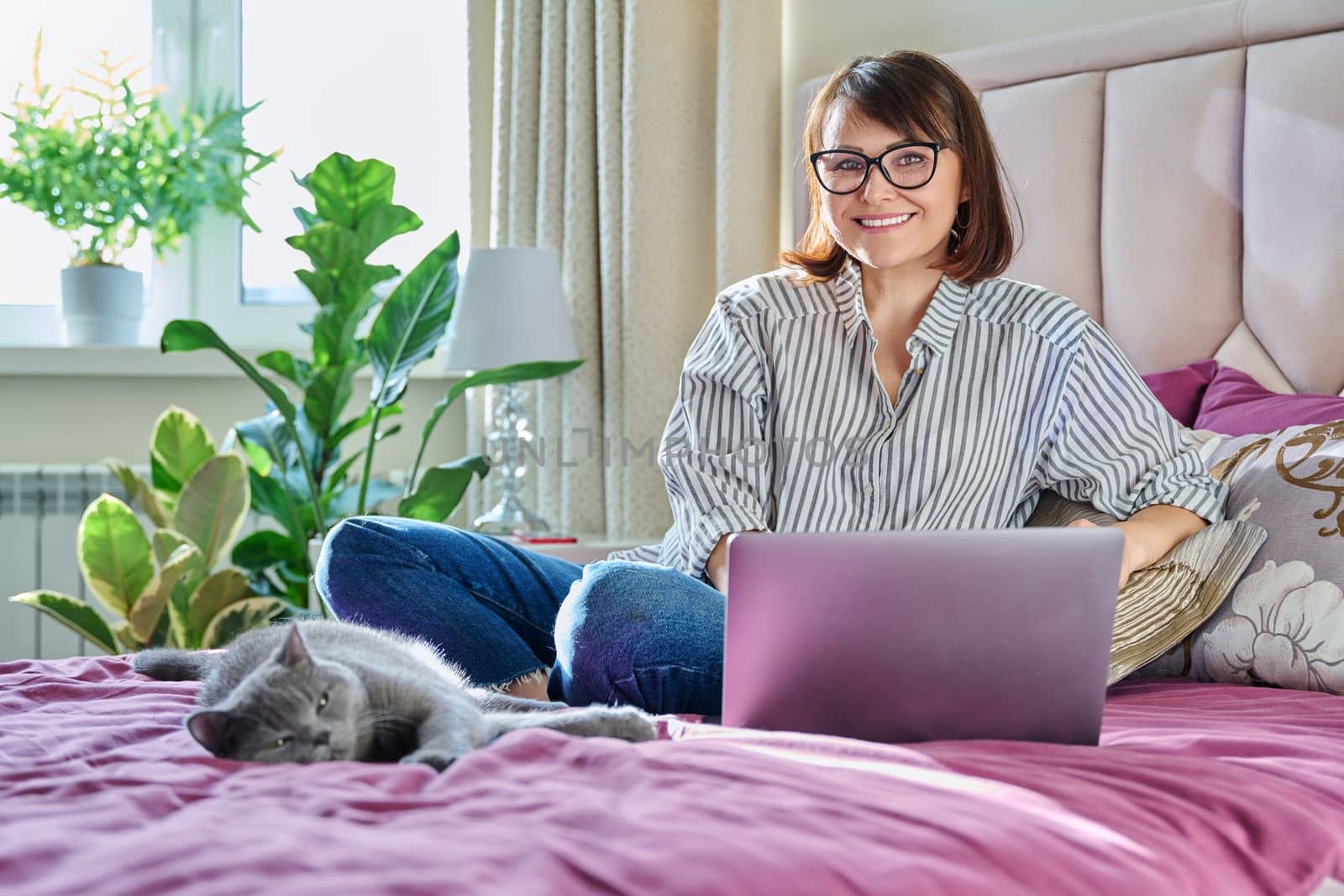 Middle aged woman at home on bed with laptop and cat. Mature 40s female looking at camera, on sofa with pet. Work at home, technology, leisure, freelancing, lifestyle, people and animals concept