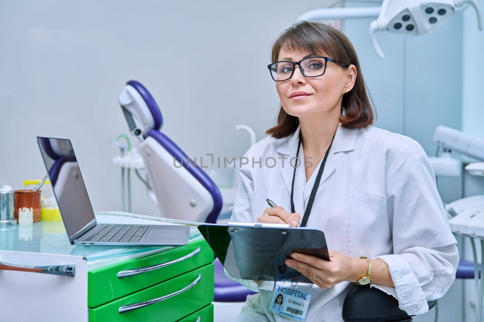 Doctor dentist working in office, looking at camera, using laptop, making notes on clipboard. Dentistry, medicine, hygiene, treatment, dental health care concept