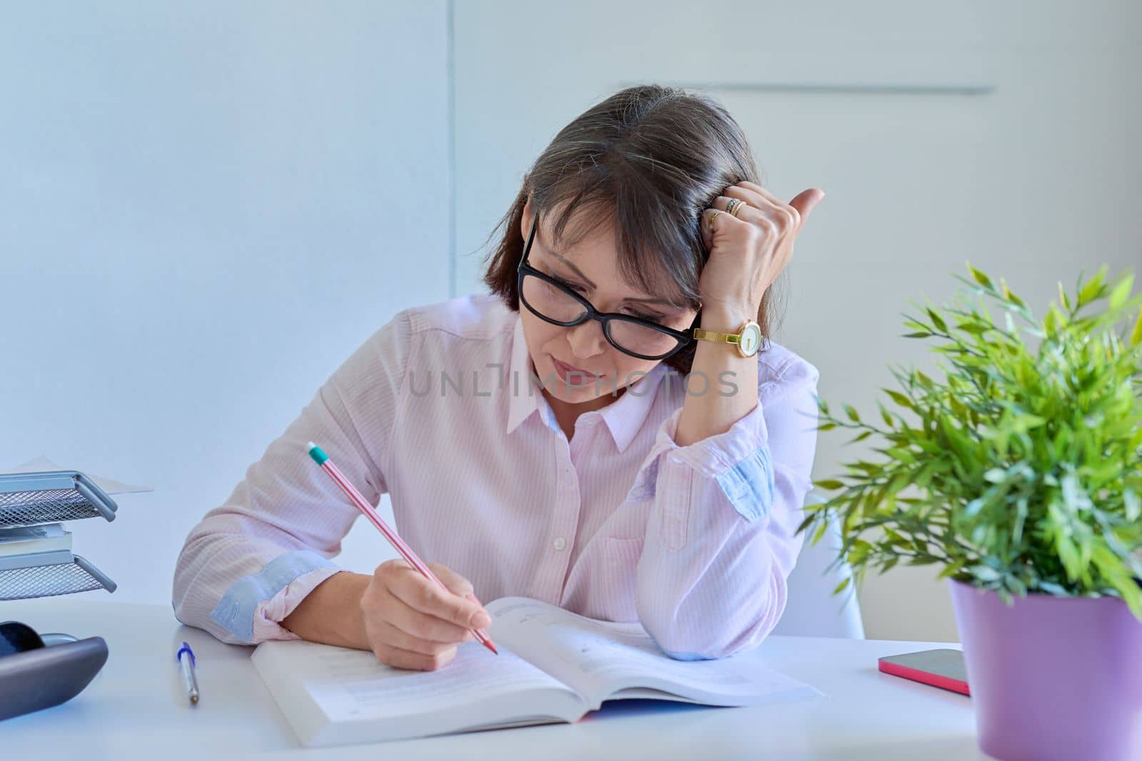 Middle-aged female working at home office at table with books. Mature woman in glasses at home workplace, education, business concept