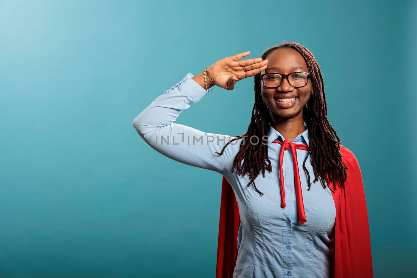 Brave and proud happy smiling superhero woman wearing mighty hero cape while making soldier salute at camera. Patriotic justice defender saluting with respect while standing on blue background.