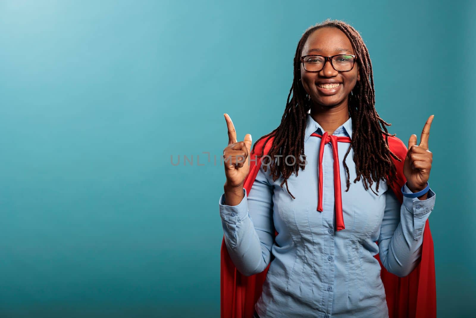 Confident and strong superhero woman wearing red cloak poiting fingers up while standing on blue background. Selfless brave justice defender posing for camera while gesturing fingers up. Studio shot