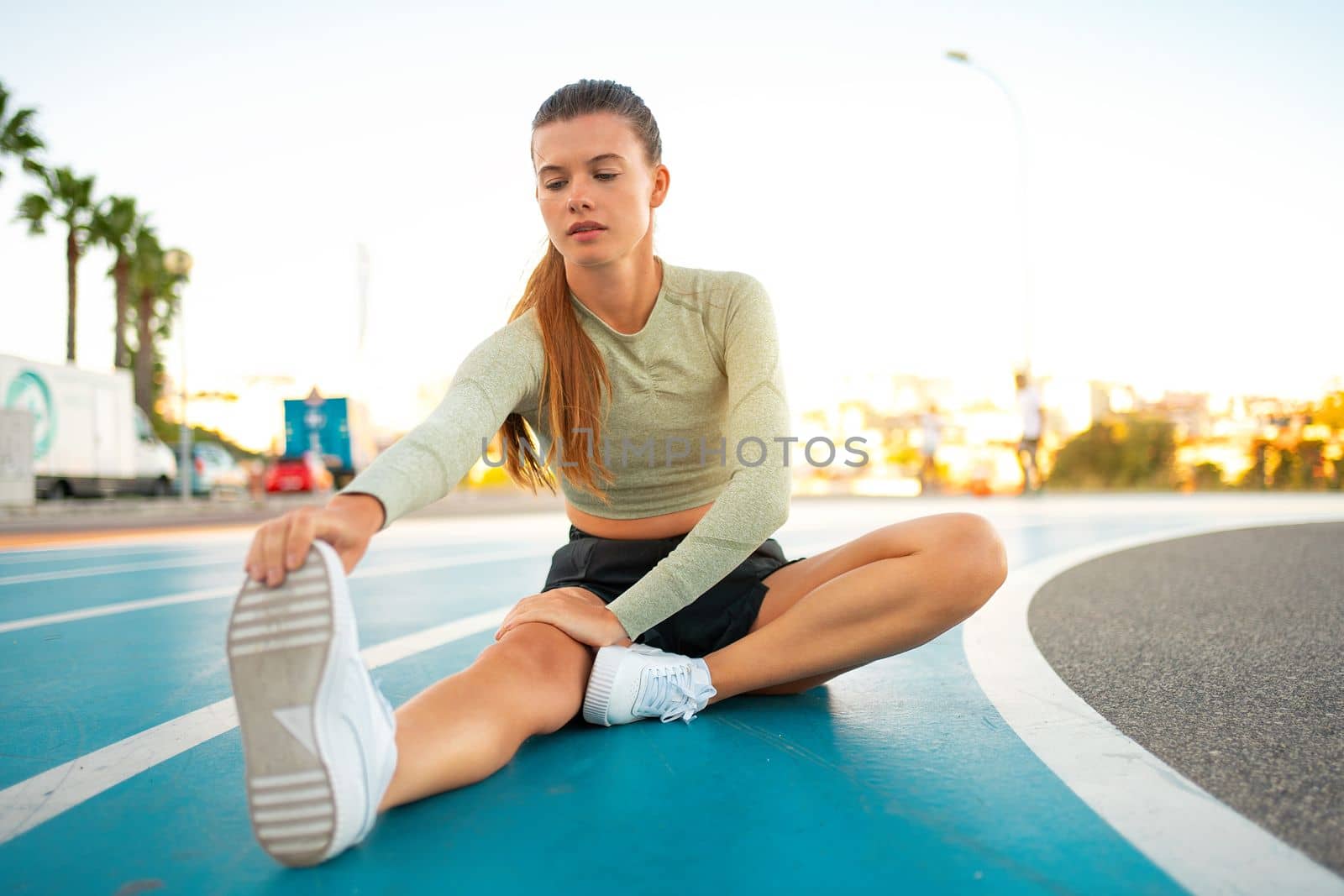 Sportswoman sitting and stretching on blue track field. Woman runner stretching leg muscles by touching his shoes. Warming up, Female runner doing stretching exercises sitting on running track.