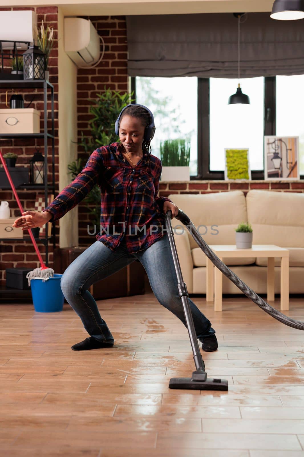 Positive housewife enjoying spring cleaning at home, singing while vacuuming in living room. Listening to music with wireless headphones relaxed looking cheerful, taking care of finishing household chores.