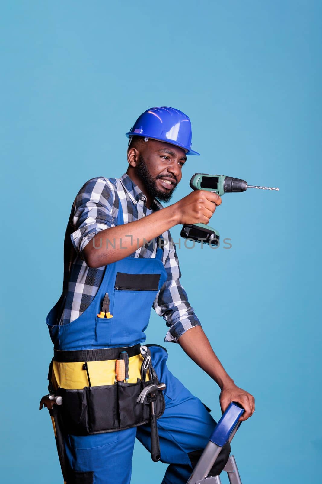 Construction worker on ladder drilling wall with cordless drill wearing work uniform and hard hat. African american man in studio shot against blue background, construction and renovation concept.