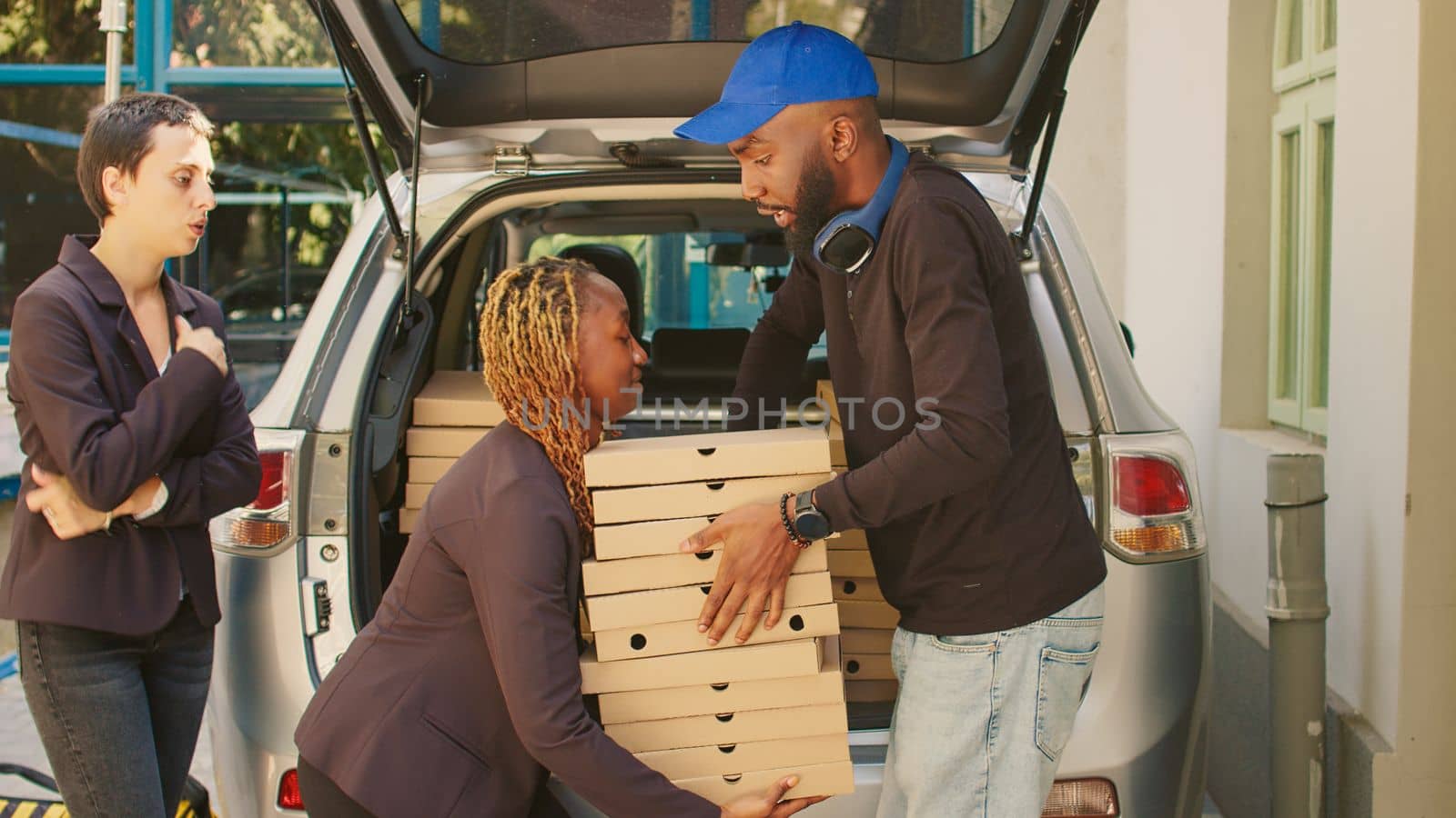Male pizzeria courier giving pizza boxes to customers at office building, delivering big restaurant order in car trunk. Taking fastfood meal packages out of vehicle to deliver to diverse women.