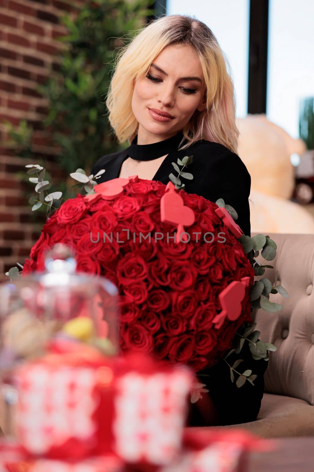 Smiling woman holding red roses bouquet, blurred giftboxes on table, valentines day luxury presents, romantic date. Excited blonde girlfriend sitting with flowers from boyfriend